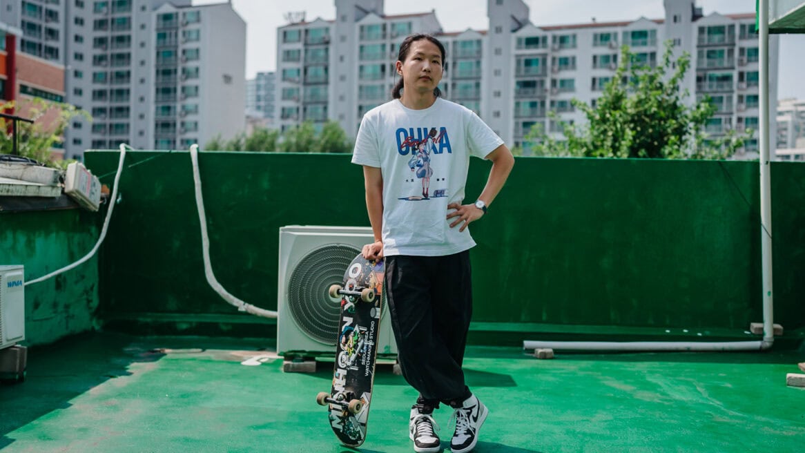 South Korean watchmaker Minhoon Yoo looks at the camera in photographer Jun Michael Park's direction in a portrait setting on a rooftop covered with waterproof green paint — very Korean. He is standing with his right hand on a skateboard, leaning on it. He is looking to his left, and he is wearing an anime T-shirt and basketball sneakers.