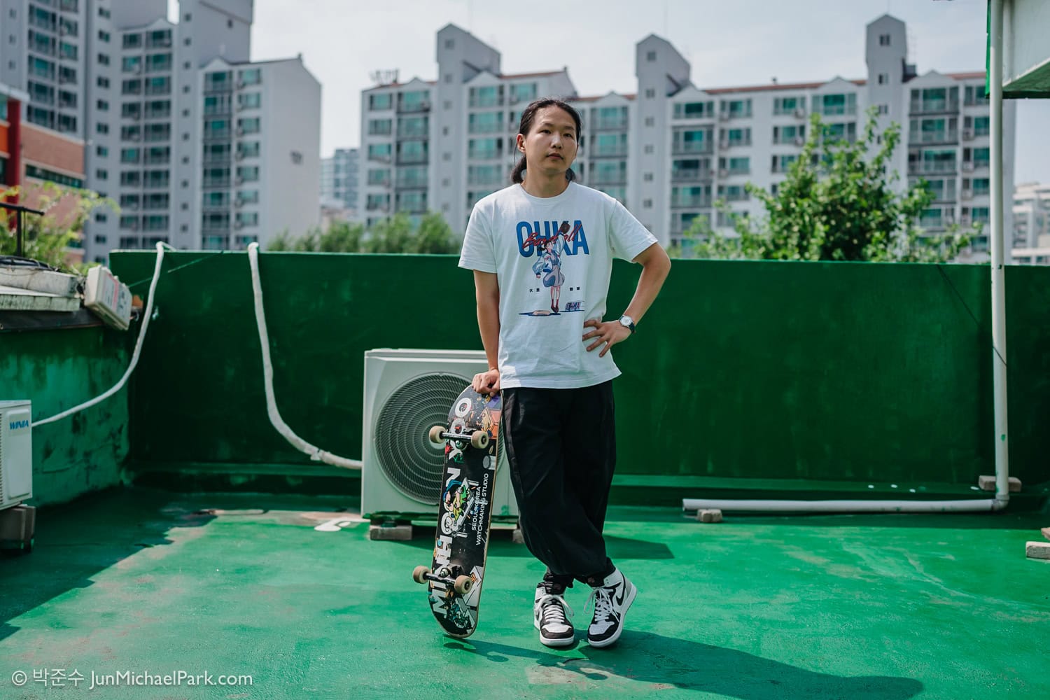 South Korean watchmaker Minhoon Yoo looks at the camera in photographer Jun Michael Park's direction in a portrait setting on a rooftop covered with waterproof green paint — very Korean. He is standing with his right hand on a skateboard, leaning on it. He is looking to his left, and he is wearing an anime T-shirt and basketball sneakers.