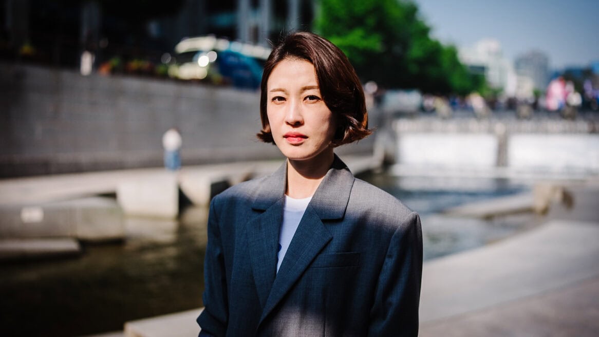 South Korean politician Ahn Gwi-ryeong, wearing a gray suit, is being photographed at Cheonggyecheon Stream by photographer Jun Michael Park. He upper body is showing in the photo, and her face is lit by sunlight while shadow is cast on part of her body. The background is blurred with a fast lens.