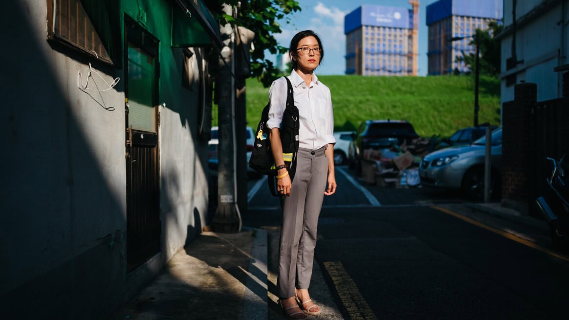 South Korean novelist Bora Chung looks at the camera at Seoul photographer Jun Michael Park's direction for a portrait. She stands in front of a shoddy house. Behind her, new apartment blocks are being built, showing a stark disparity between the rich and poor.