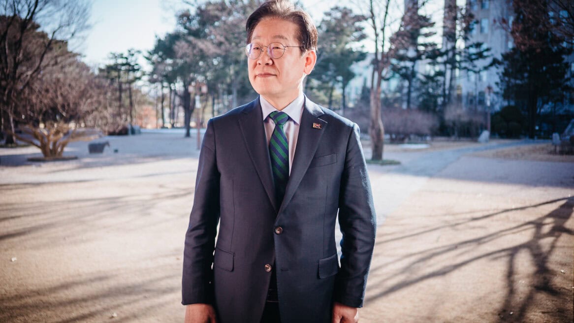 South Korean politician Lee Jae-myung, wearing a dark suit and a green-black-stripe tie, is directed to look to his right during a portrait session with Seoul photographer Jun Michael Park at the National Assembly. The background shows well-grown trees and their shadows, as well as part of the main hall.